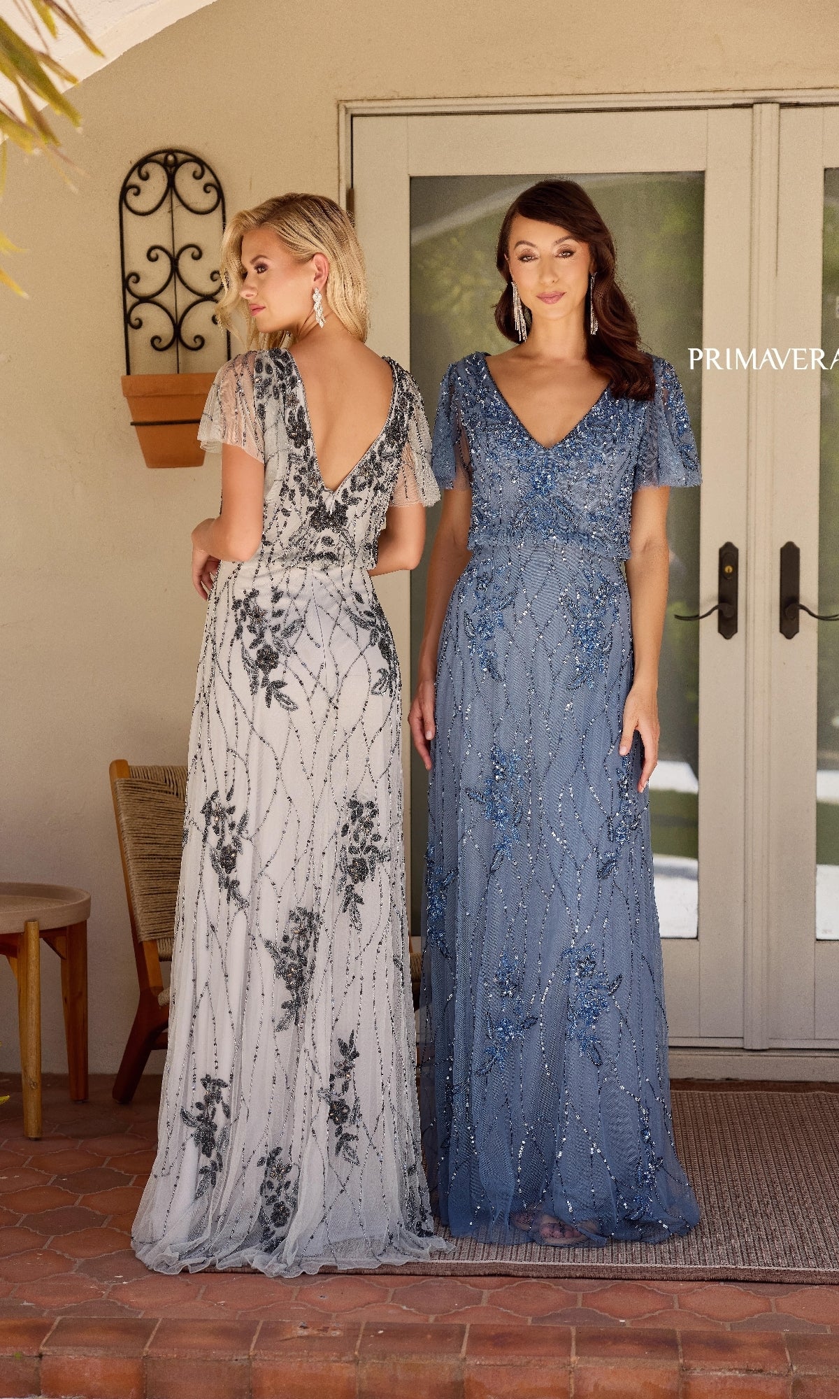 Two women stand before double doors, both in floor-length, short-sleeved beaded gowns. One wears the silver-gray Primavera 13144 formal A-line dress with black details, the other is dressed in blue.